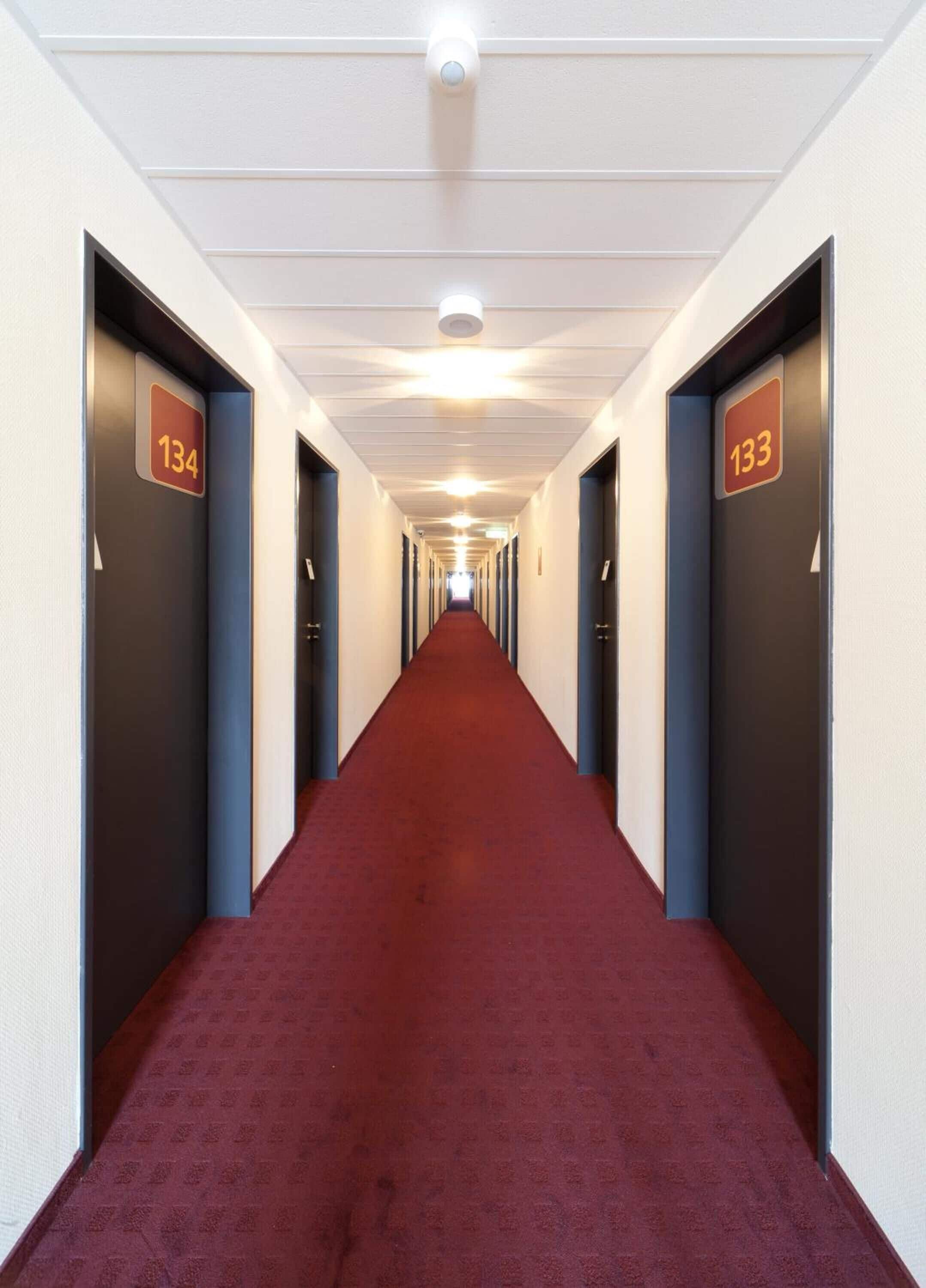 Pasillo de hotel con alfombra roja, puertas numeradas y una lámpara en el techo.