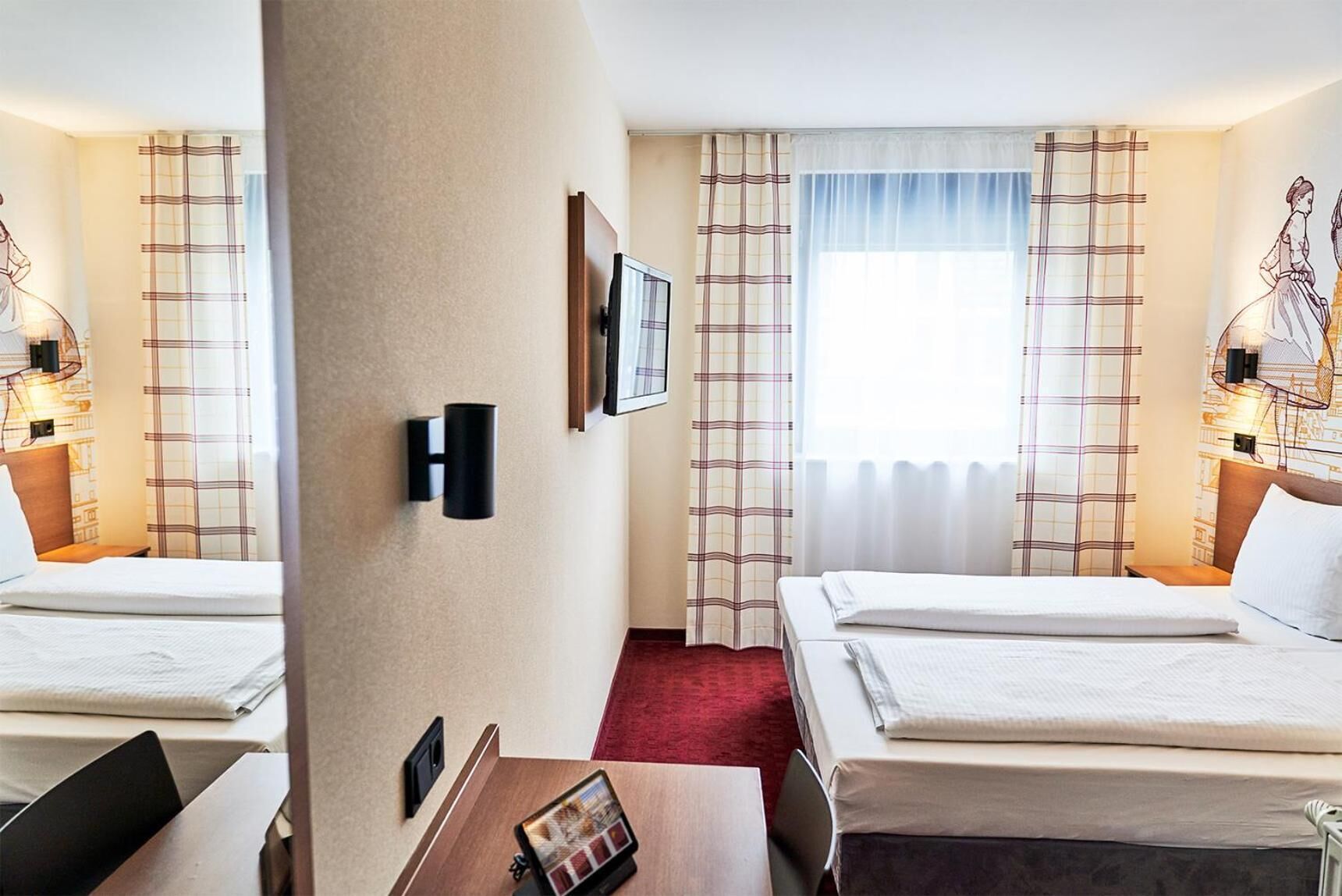 A hotel room with two beds, a red carpet, a wall-mounted TV, and a window with plaid curtains.