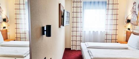 A hotel room with two beds, a red carpet, a wall-mounted TV, and a window with plaid curtains.