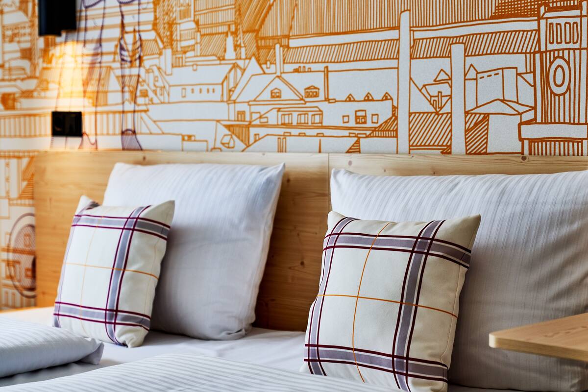 A hotel room with a bed, two pillows, a wooden headboard, and a wall with a cityscape mural.