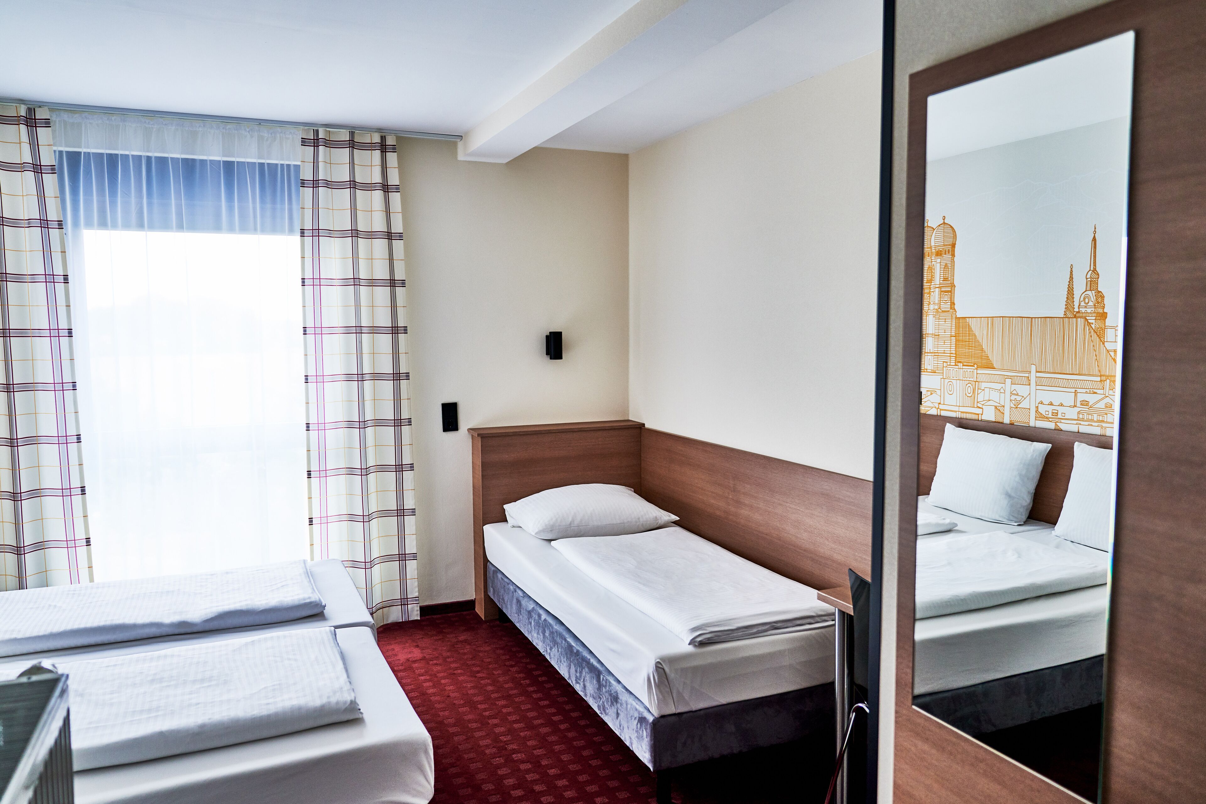 Habitación de hotel con dos camas, alfombra roja, ventana con cortinas y un espejo que refleja la habitación.