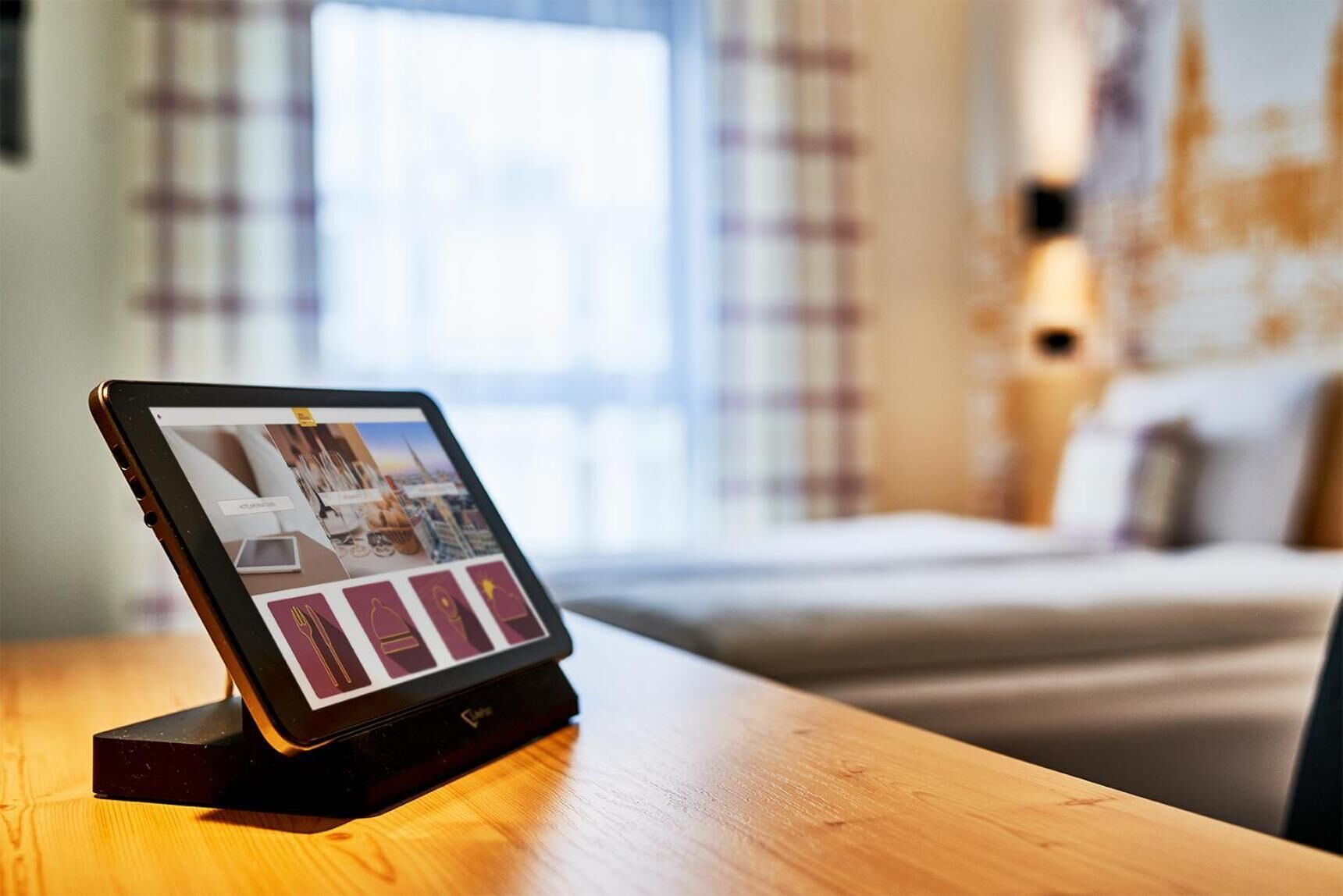 Une tablette posée sur une table en bois affiche un plan d’une chambre d’hôtel, avec un lit, une fenêtre avec des rideaux et un mur muni d’un luminaire.