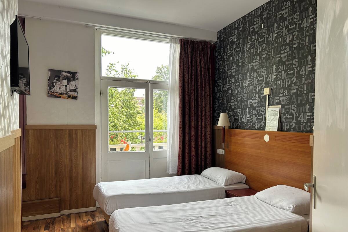 A hotel room with two beds, a window with curtains, a wooden floor, a wall with a pattern, and a mounted TV.