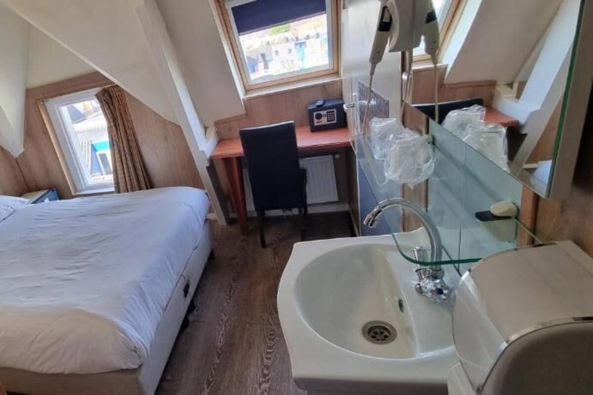 A compact bedroom with a single bed, a small desk, a mirror, and a sink.