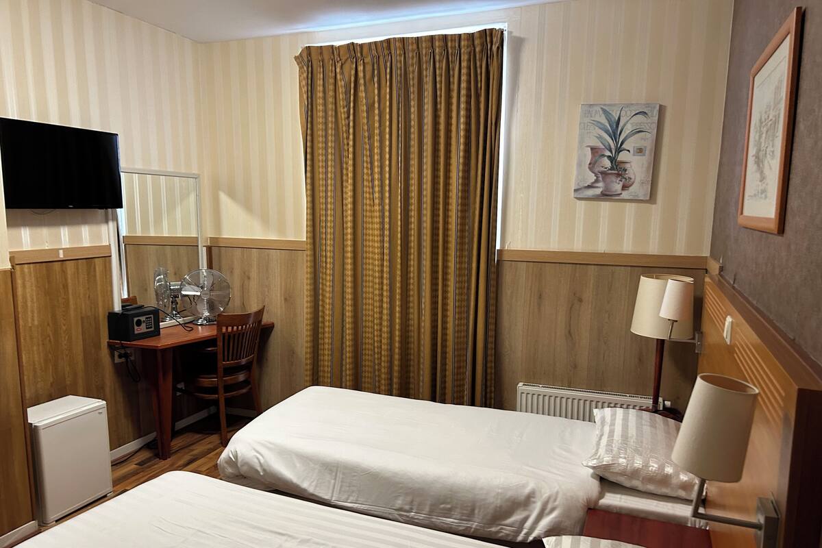A hotel room with two beds, a desk with a chair, a television, a radiator, and a lamp.