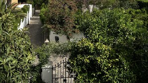 Un estrecho sendero empedrado bordeado de abundante vegetación y altos cipreses, que conduce a un edificio blanco con tejado de baldosas.