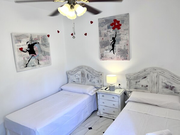 Quarto com duas camas, mesa de cabeceira com abajur e duas obras de arte na parede retratando figuras com corações.