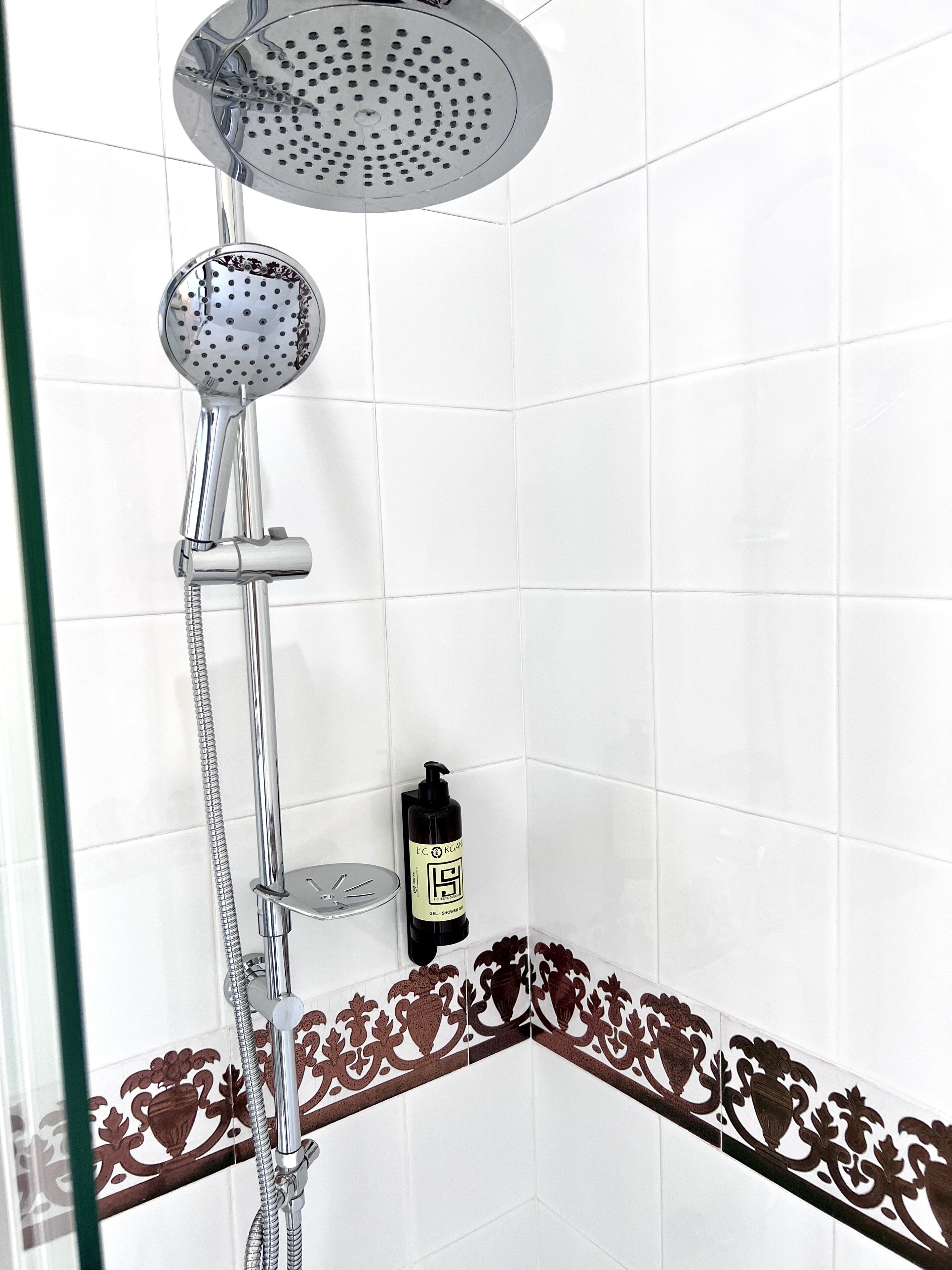 A shower with a handheld showerhead and a wall-mounted showerhead, a glass shelf with a bottle of shampoo, and decorative tiles with a brown pattern.