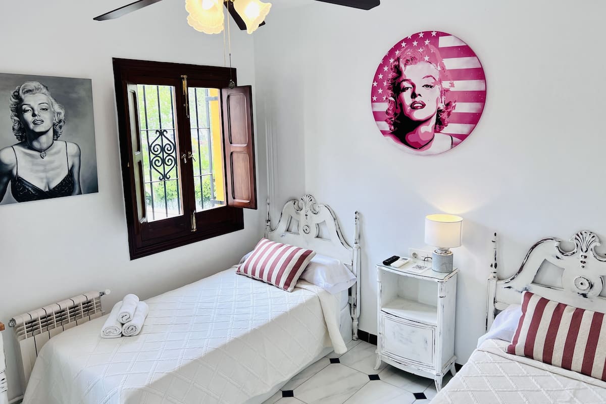 A bedroom with two beds, a ceiling fan, a window with curtains, and artwork on the walls.
