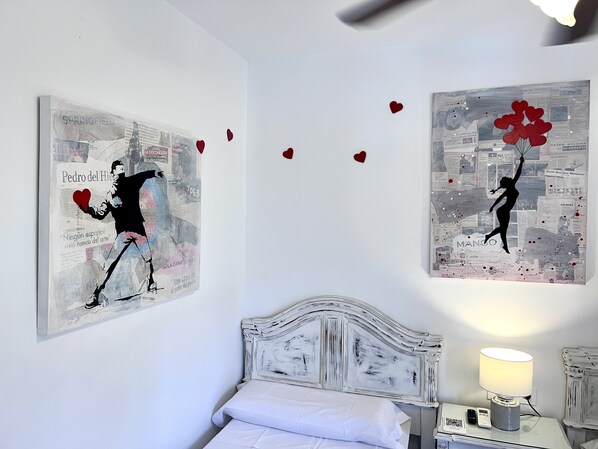 Un dormitorio con una cama blanca, un cabecero blanco y dos obras de arte en la pared que muestran figuras con corazones.