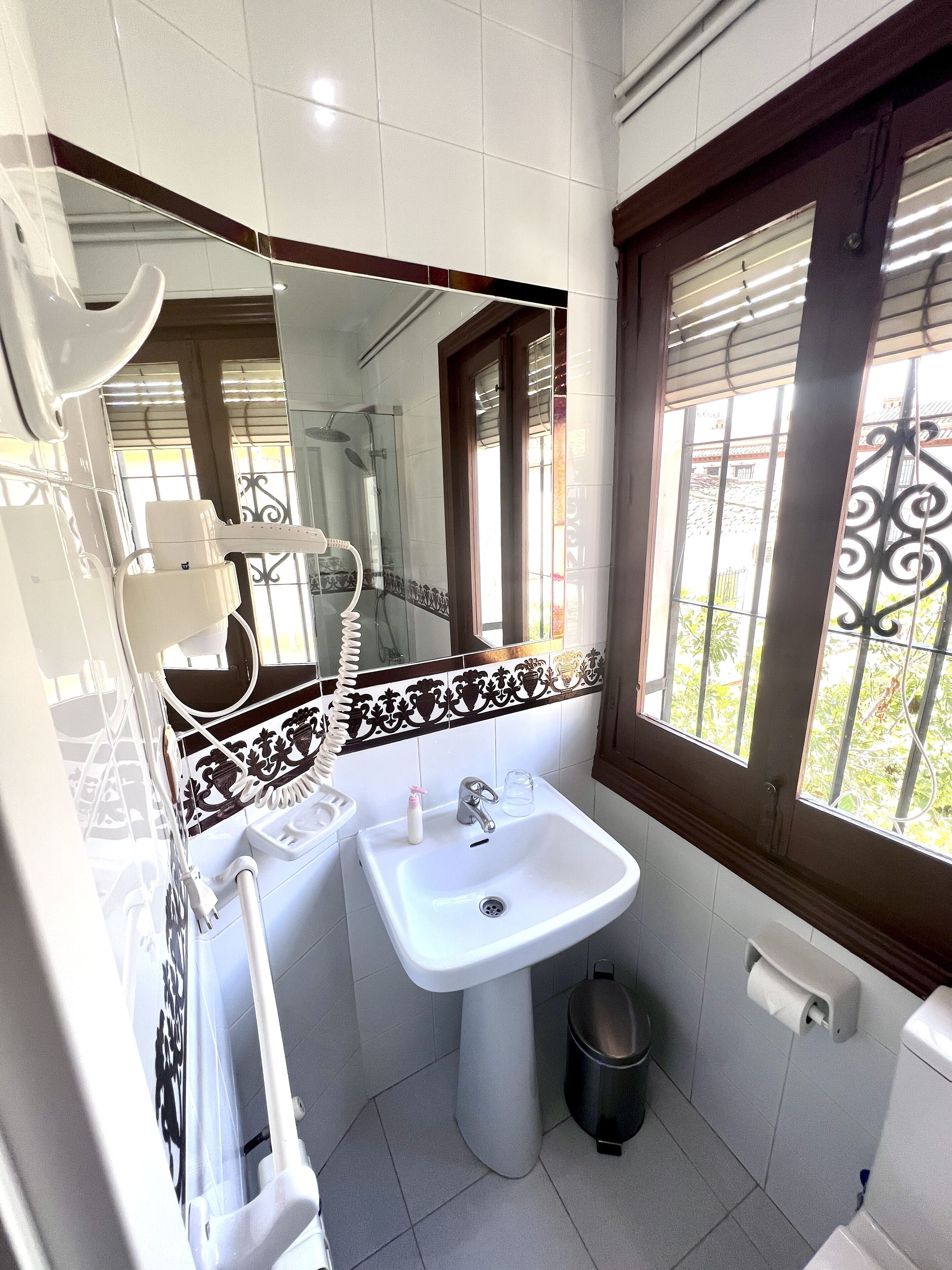 Un baño con un lavamanos blanco, un espejo, una ventana con una reja de hierro decorativa y un cesto de basura.