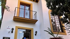 Un edificio de dos plantas con fachada amarilla, un balcón con barandilla de hierro forjado y una planta grande en maceta al frente.