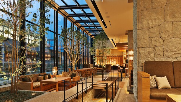 A modern hotel lobby with a glass ceiling, indoor plants, and a stone wall.
