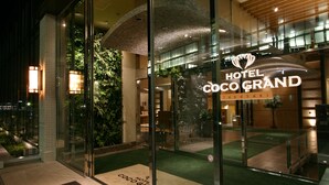 The entrance of Hotel Coco Grand with a glass facade, indoor plants, and a visible staircase leading to the upper floor.
