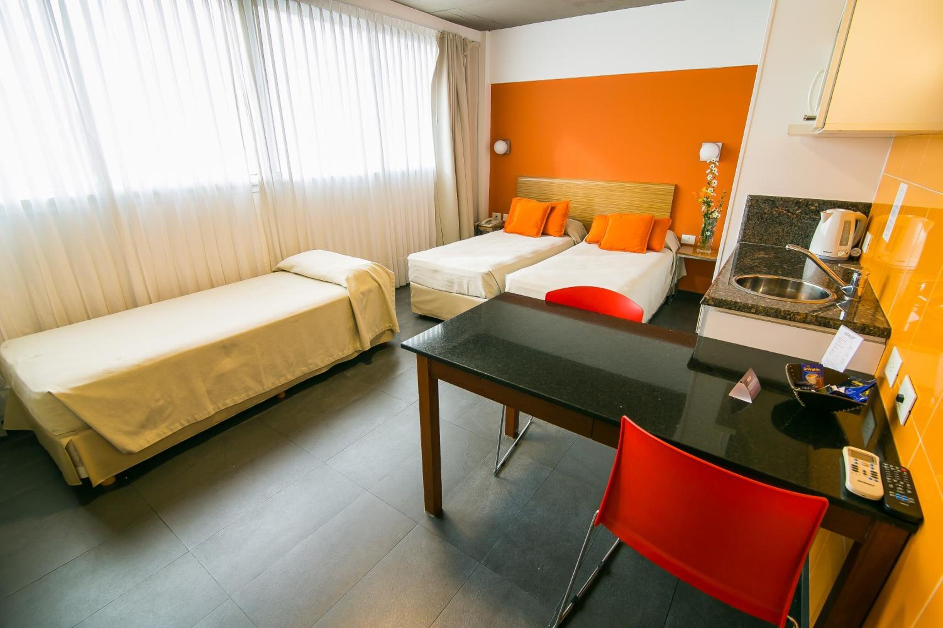 A hotel room with two single beds, a small kitchenette, a dining table, and a red chair.