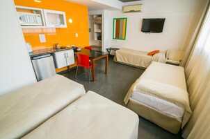 Habitación de hotel pequeña con dos camas individuales, una kitchenette, una mesa de comedor con una silla roja, un televisor y un espejo.