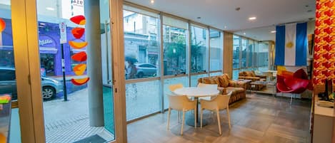 A modern hotel lobby with a glass entrance, a seating area with a round table and chairs, and a view of the street outside.