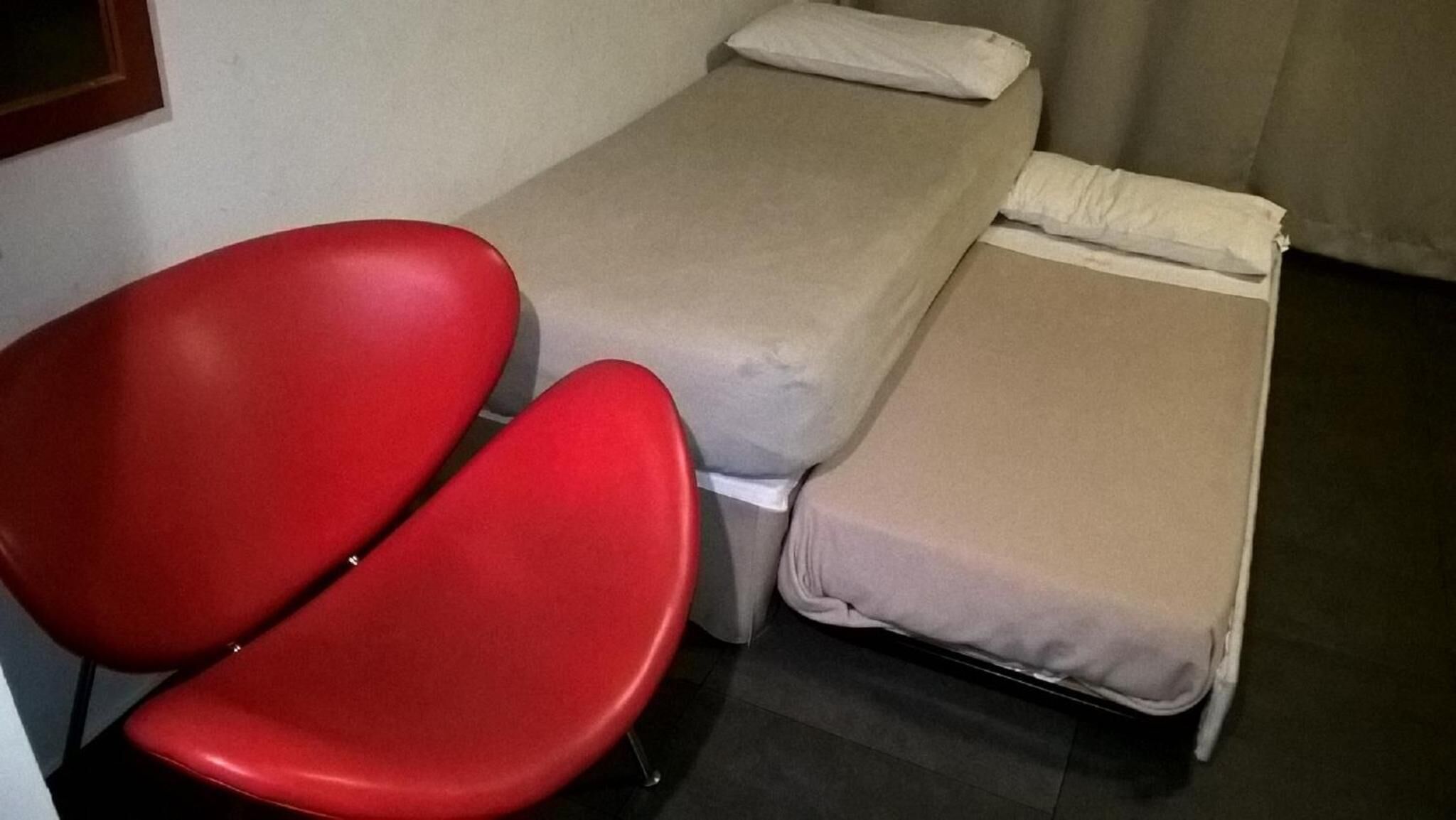 Una habitación pequeña con una cama individual, una silla roja y una mesita.
