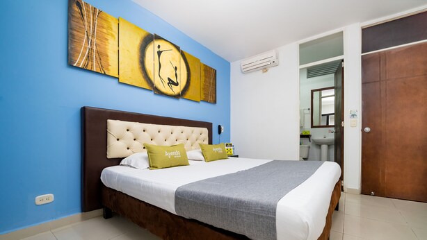 A hotel room with a large bed, a headboard, a wall-mounted artwork, an air conditioning unit, a bathroom visible through an open door, and a brown door. - Ayenda 1703 Gran Reserva (Villavicencio)