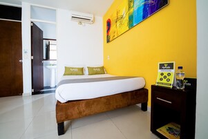 A hotel room with a bed, a bedside table with a water bottle, a sign with room amenities, and a colorful abstract painting on the wall.