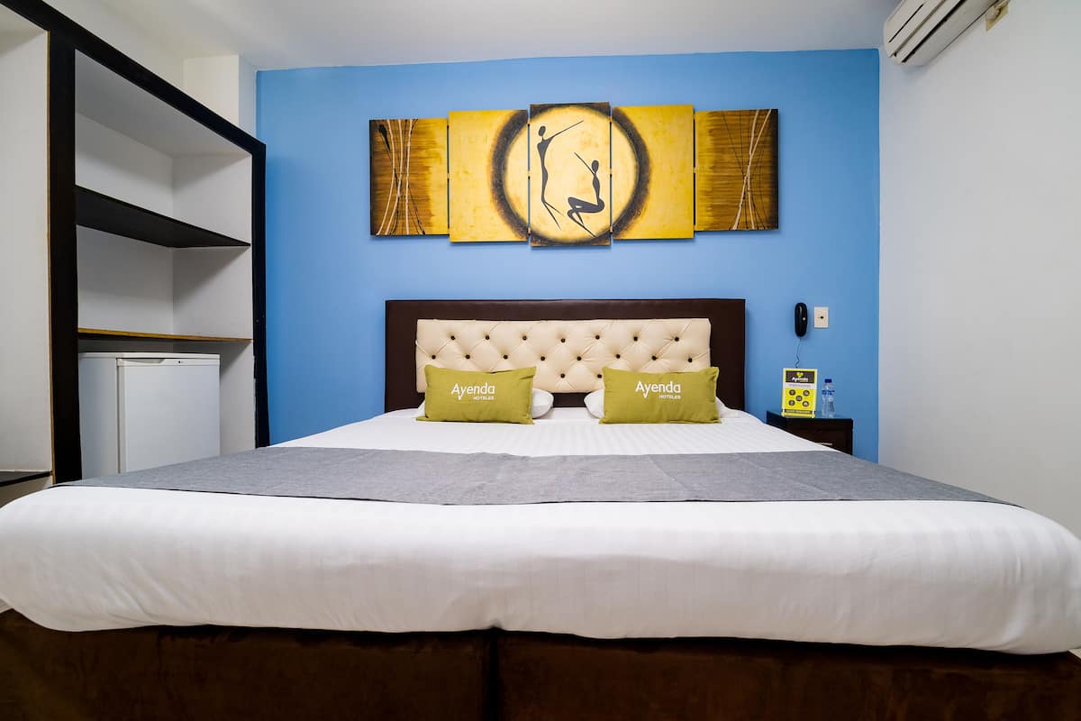 A hotel room with a large bed, a headboard with pillows, a wall art piece, a small table with a water bottle, and a power outlet.