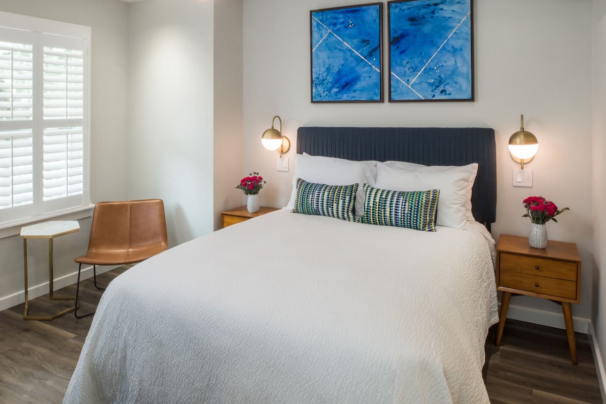 A neatly arranged bedroom with a large bed, two bedside tables with lamps, a chair, and a window with blinds.