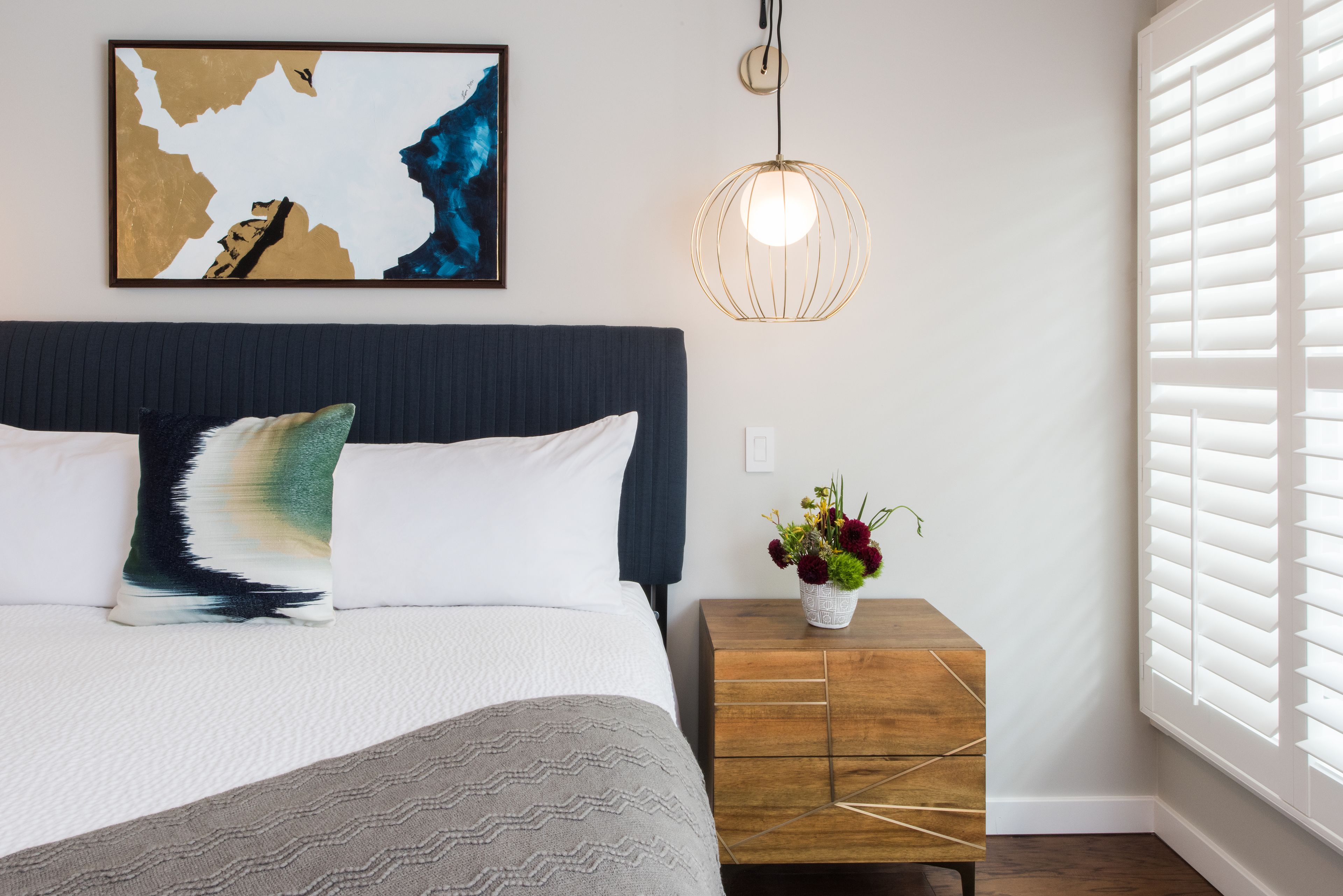 A neatly arranged bedroom with a bed, a bedside table with a vase of flowers, a hanging light fixture, and a window with blinds.