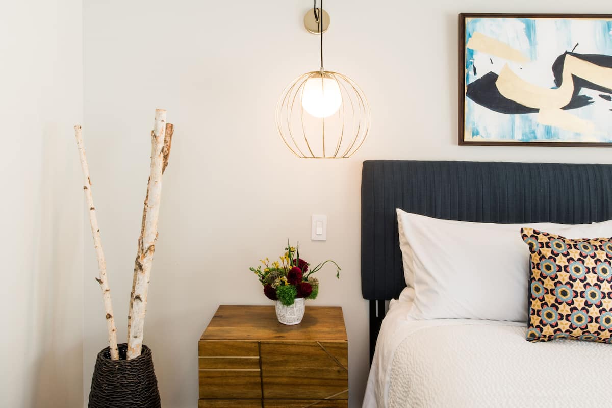 A bedroom with a bed, a bedside table with a vase of flowers, a wall-mounted light fixture, and a piece of abstract art.