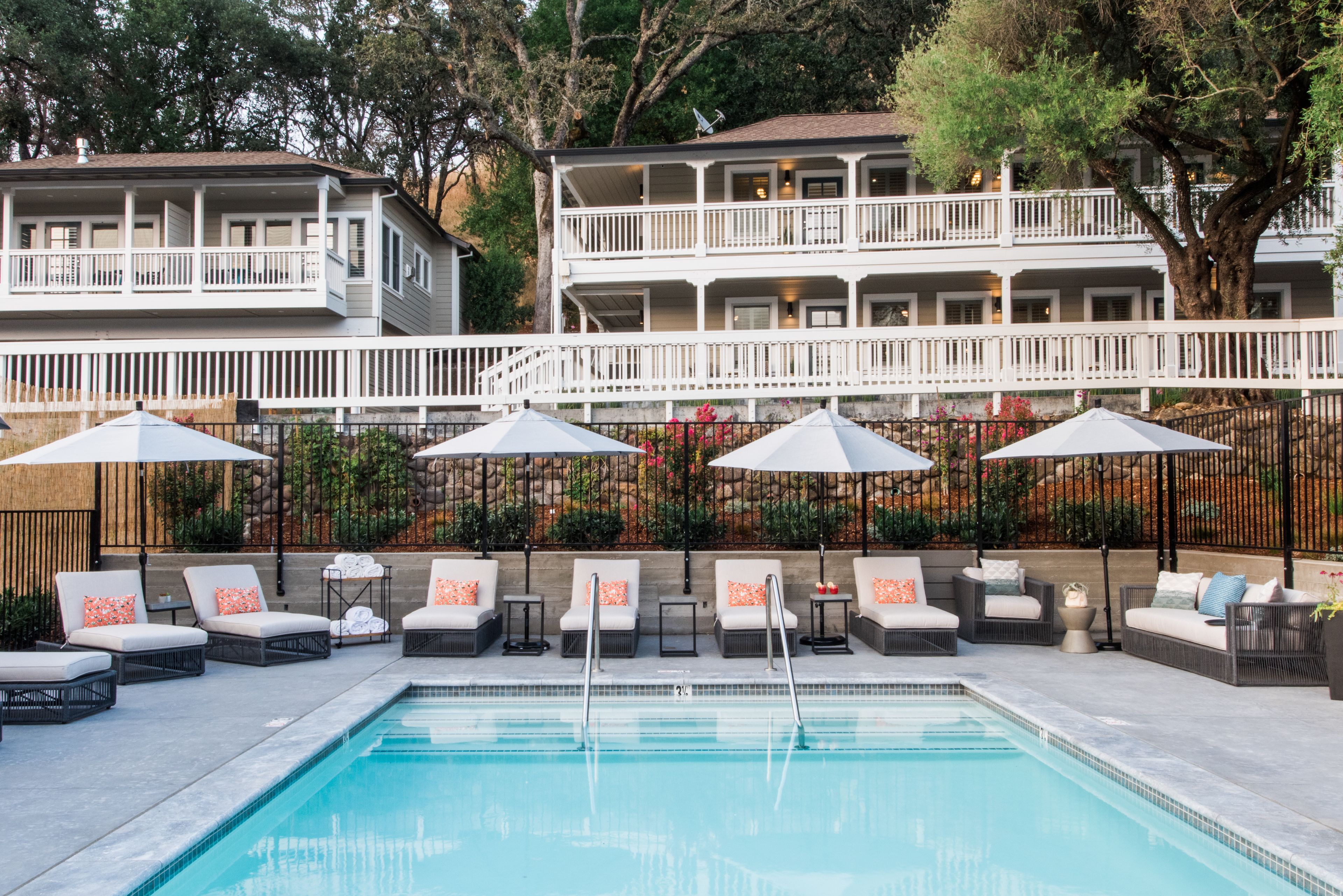 A pool area with lounge chairs and umbrellas, a white fence, and a building with balconies in the background.