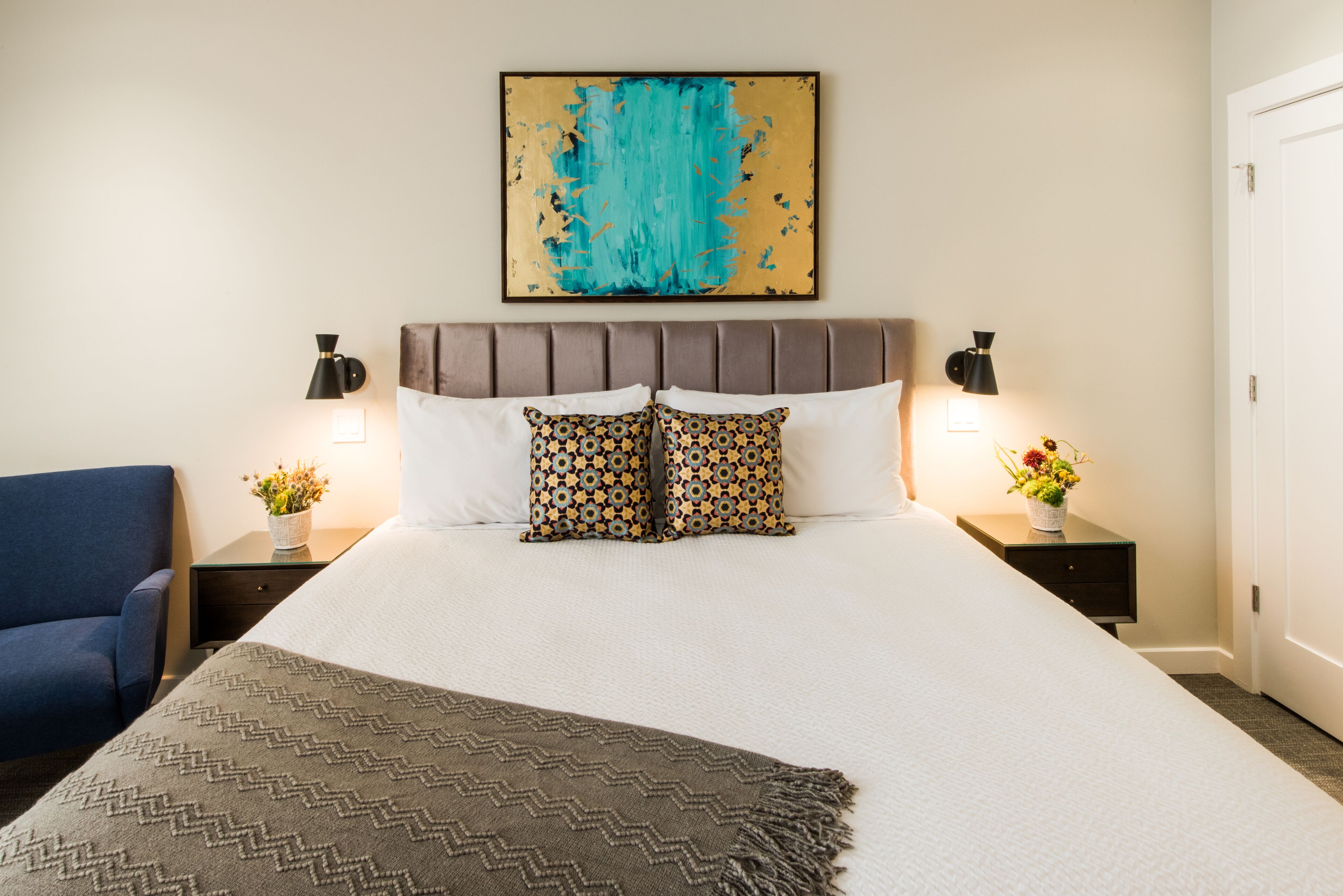 A neatly arranged hotel room with a large bed, two bedside tables with lamps, a blue armchair, and a decorative painting on the wall.