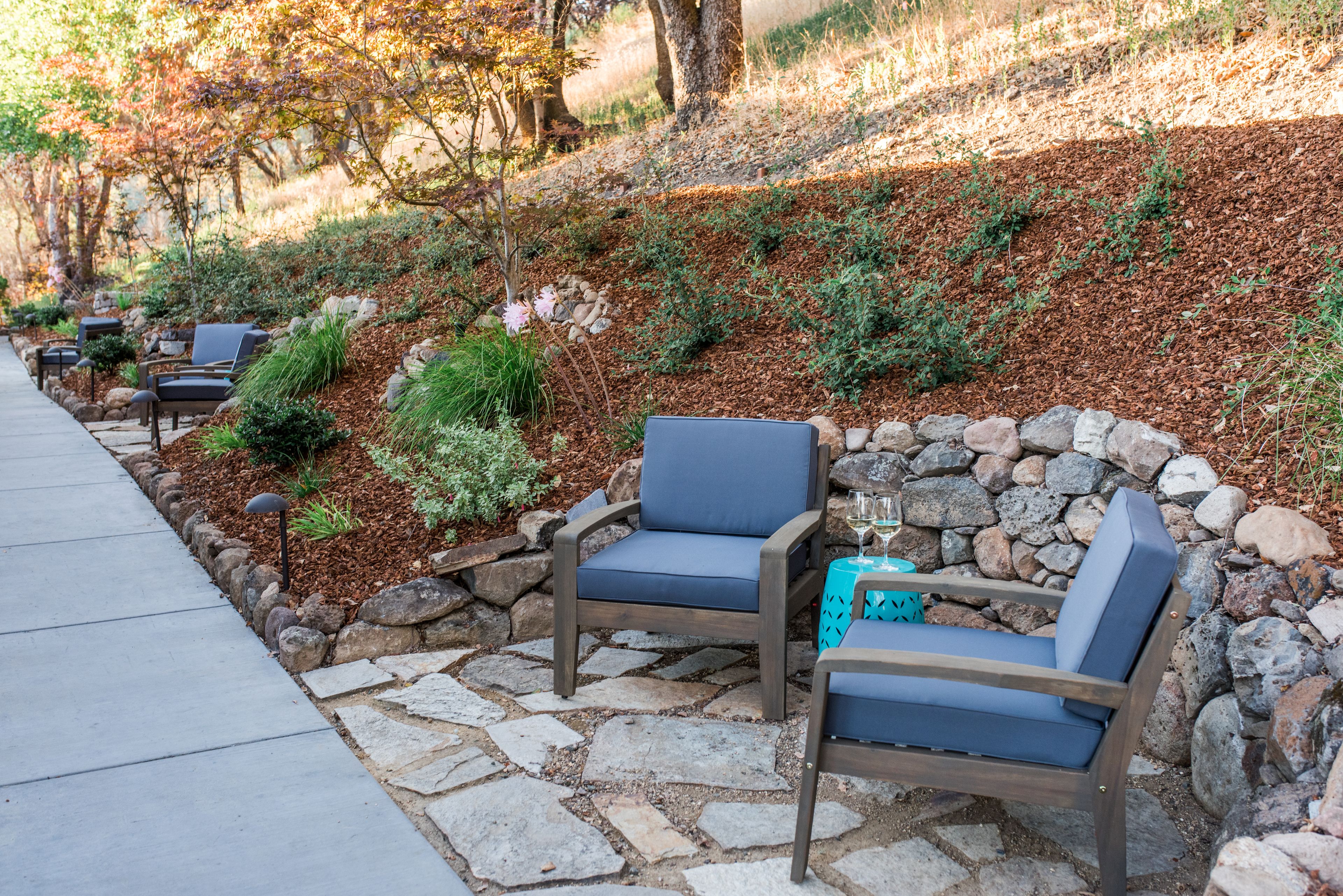 A stone pathway leading to outdoor seating with chairs and a table, surrounded by a landscaped garden with various plants and a stone wall.