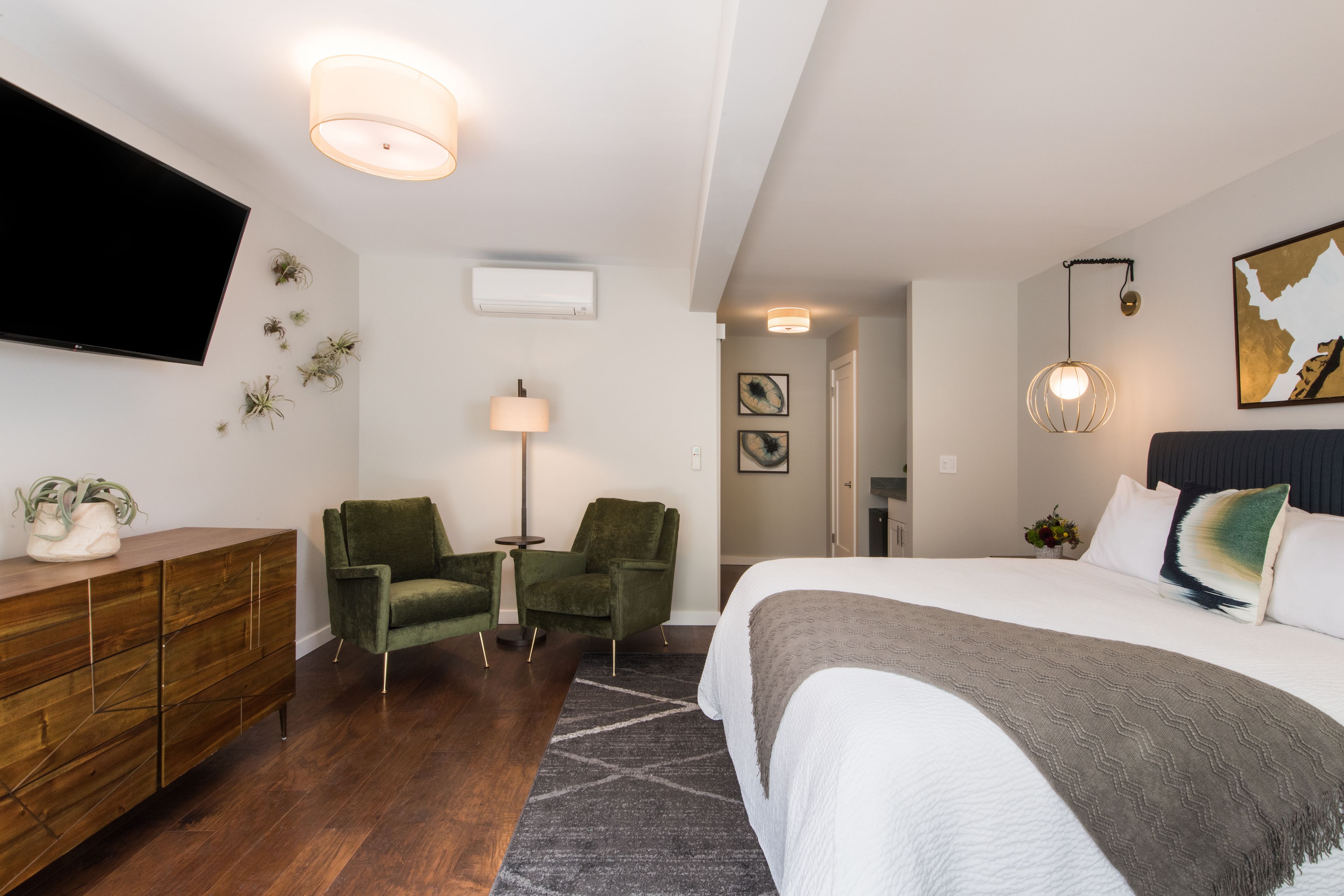 A modern hotel room with a large bed, a flat-screen TV, a wooden dresser, two green armchairs, a small table, and a floor lamp.
