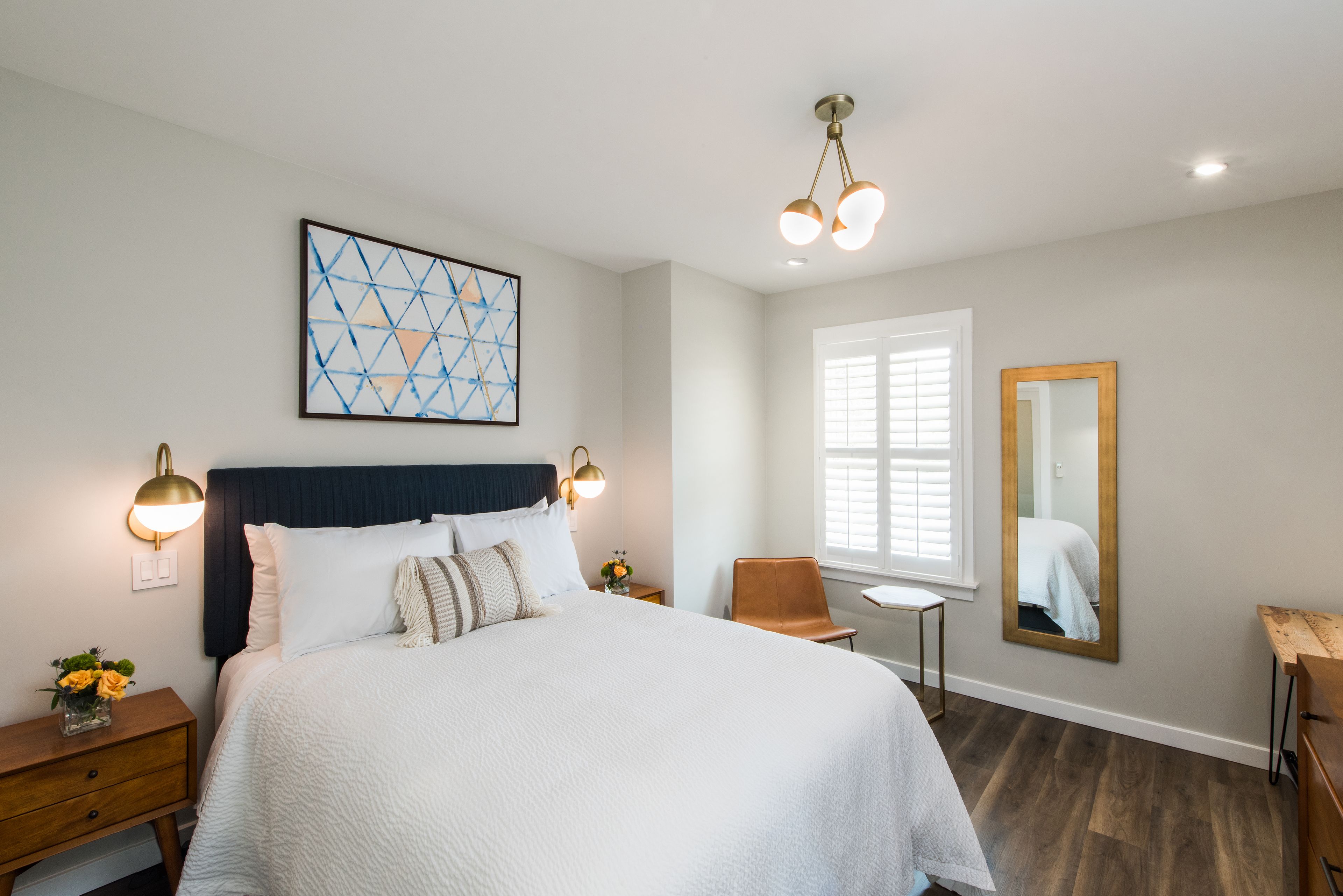 A bedroom with a large bed, a nightstand with a lamp, a chair, a small table, a window with blinds, and a mirror on the wall.