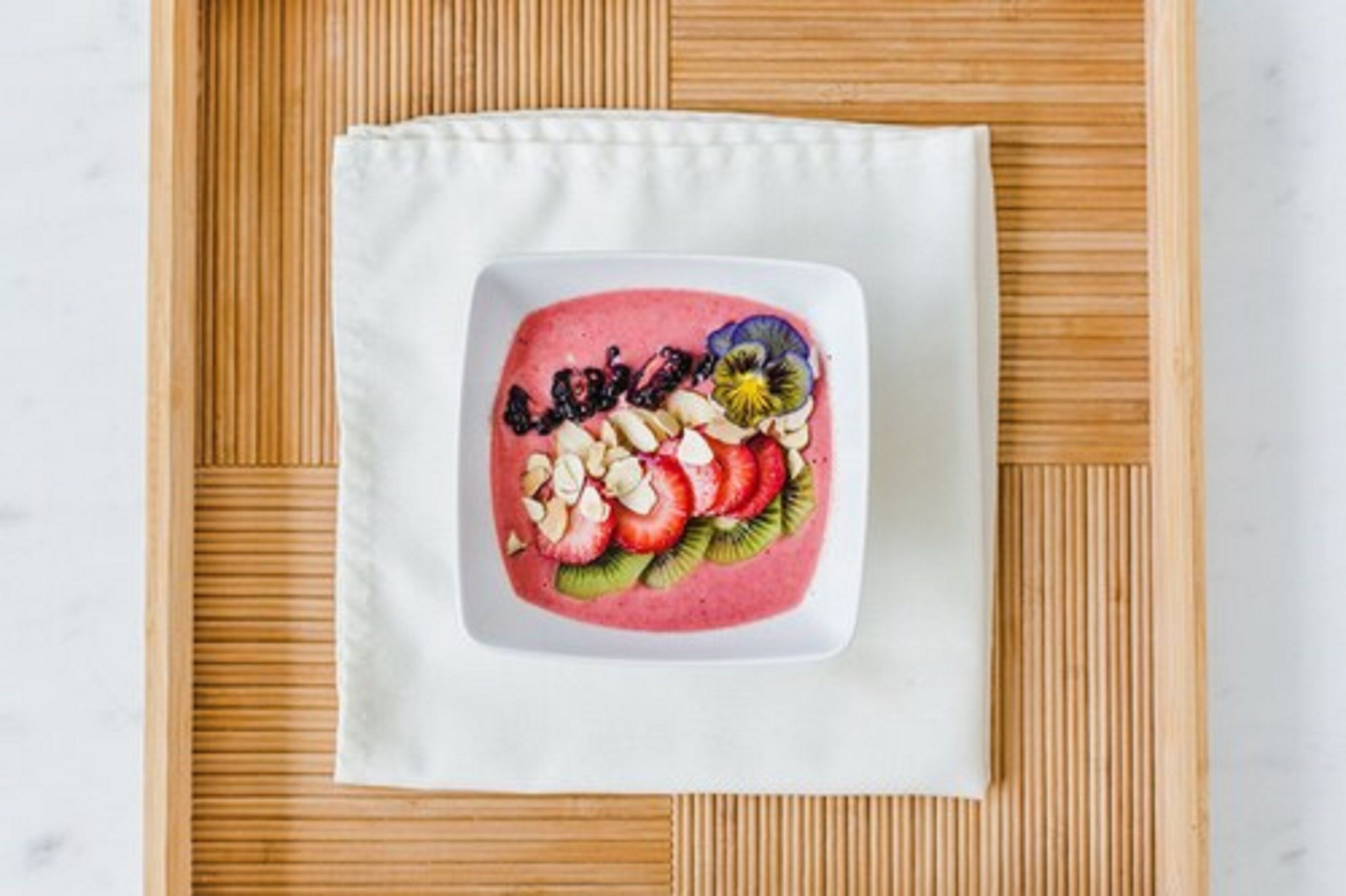 Ein quadratischer, weißer Teller mit einem rosa Smoothie, belegt mit Obstscheiben und Nüssen auf einer Bambusmatte.