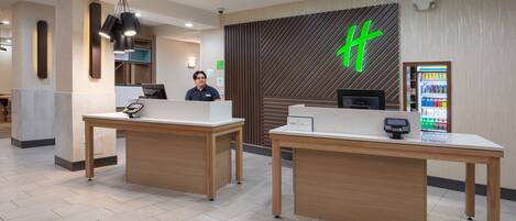 A hotel reception area with two reception desks, a digital screen, a vending machine, and a large green logo on the wall.