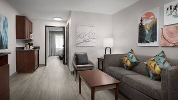 A modern living room with a sofa, a coffee table, and a dining area with a microwave. There are framed artworks on the walls and a lamp on a side table.