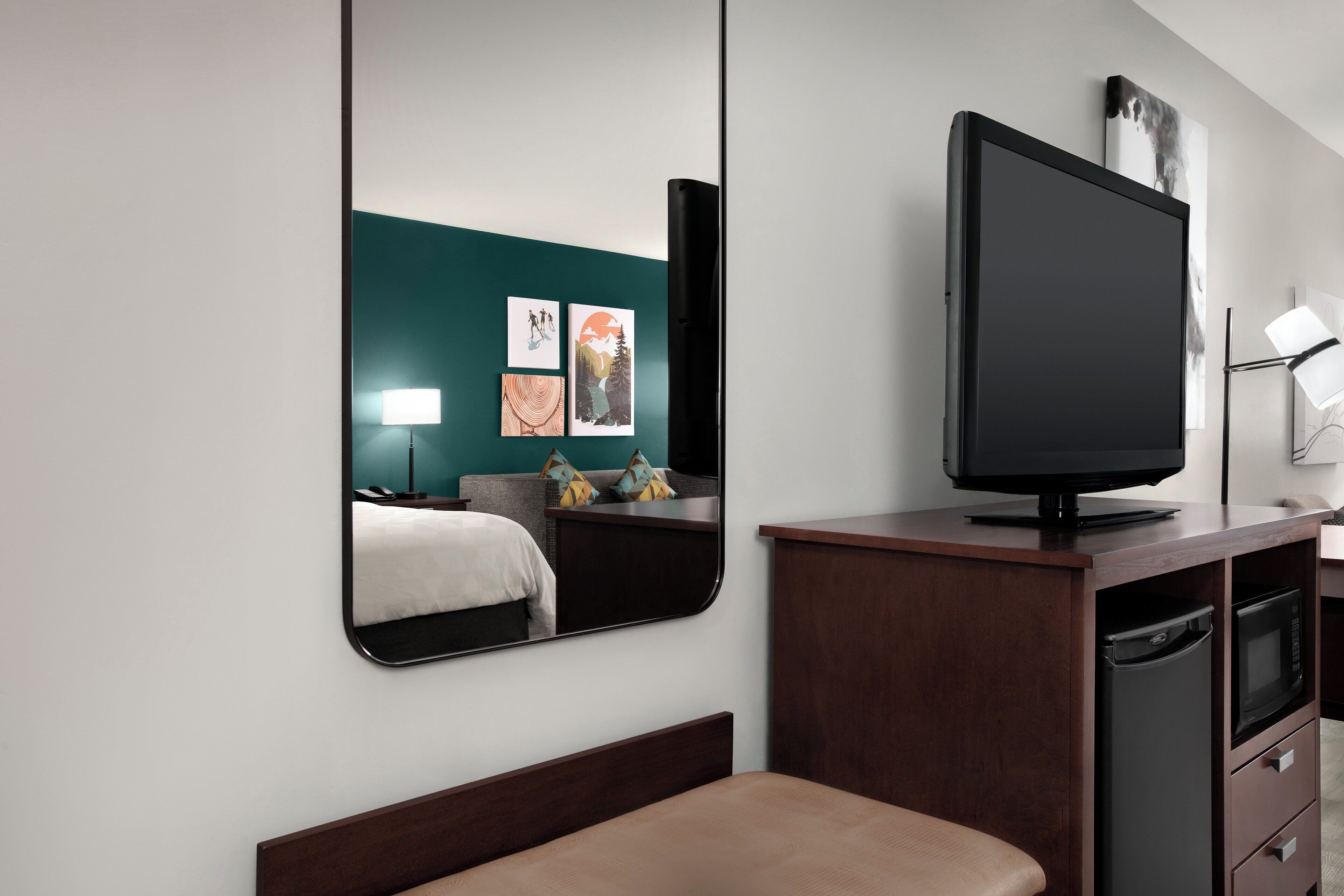 Hotellihuone, jossa on sänky, televisio, peili, lipasto ja taideteoksia seinällä.