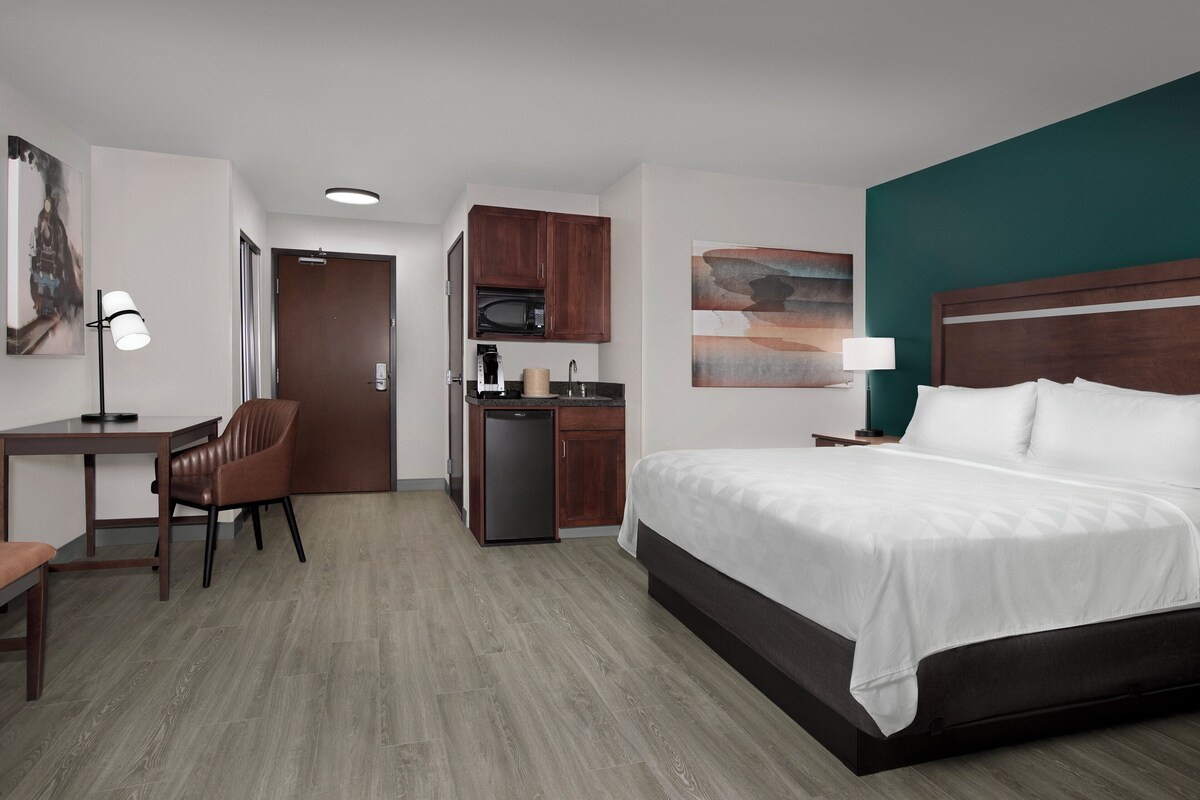 Ein Hotelzimmer mit Bett, Schreibtisch, Stuhl, Küchenzeile und Kunstwerken.