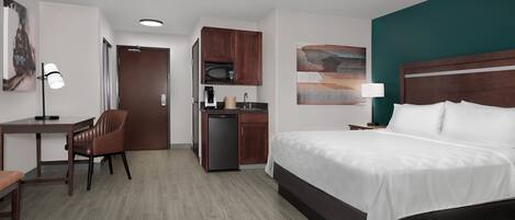 A hotel room with a bed, desk, chair, kitchenette, and artwork.