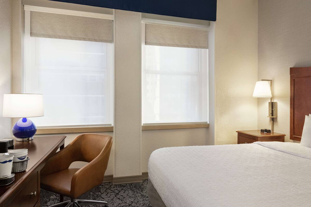 A hotel room with a bed, a desk with a lamp, a chair, and a nightstand with a lamp.