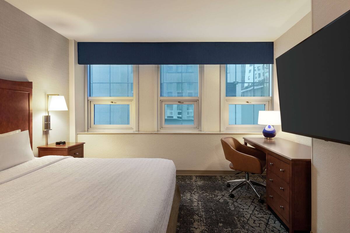 A hotel room with a large bed, two bedside tables with lamps, a desk with a chair, and a large window with a view of buildings.