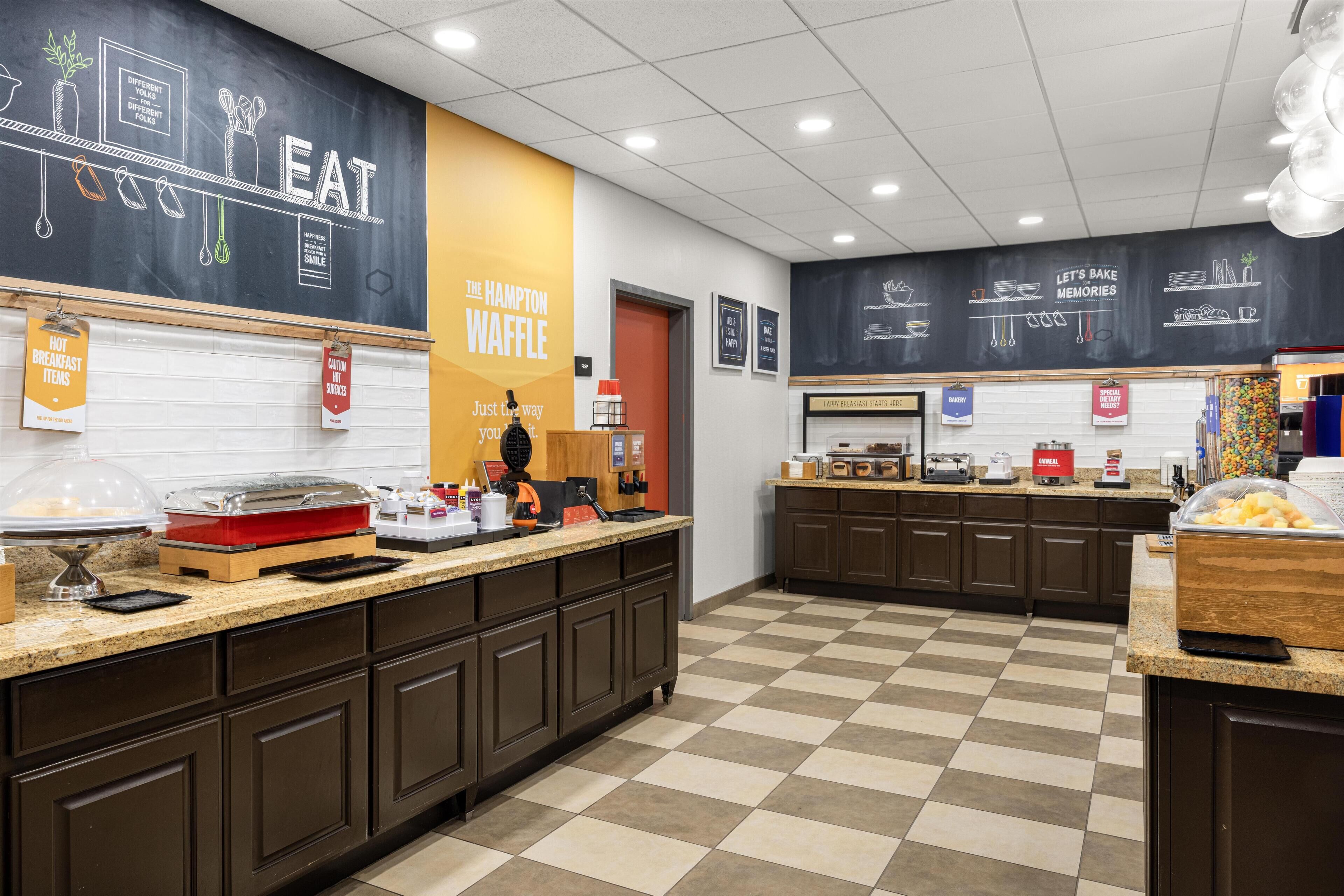 A modern kitchen with a chalkboard wall displaying food-related drawings and text, a buffet counter with various food items, and a checkered floor.