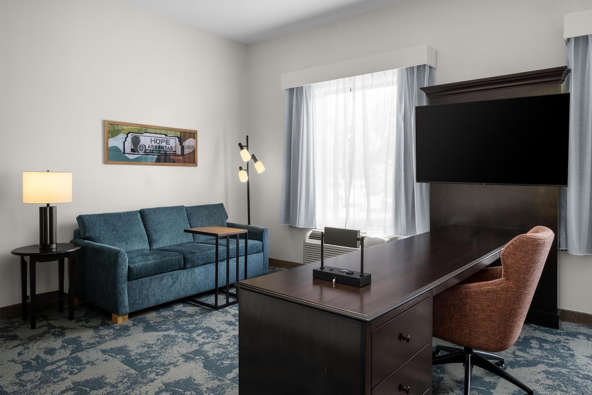 A hotel room with a dark wooden desk, a blue sofa, a small table, a lamp, a TV, and a window with curtains.