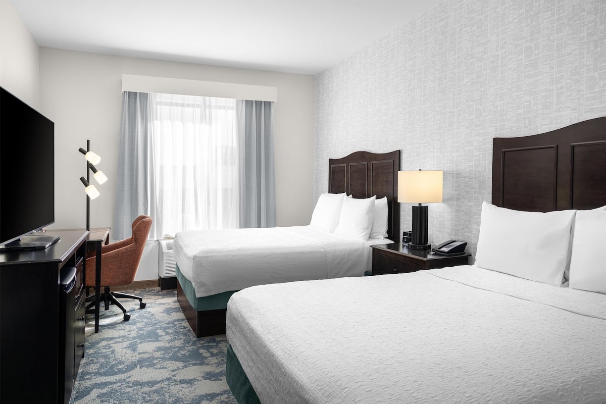 A hotel room with two beds, a desk with a TV, a chair, a lamp, and a window with curtains.