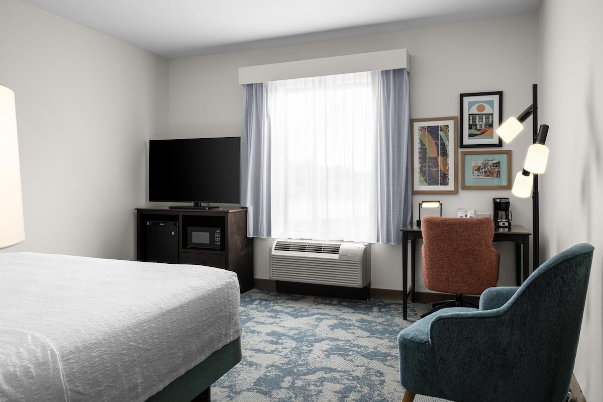 A hotel room with a bed, a desk with a computer, a chair, a TV, and a window with curtains.