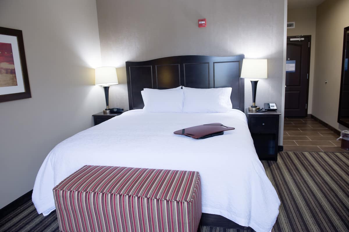 A hotel room with a large bed, two bedside tables with lamps, a striped ottoman, and a framed picture on the wall.