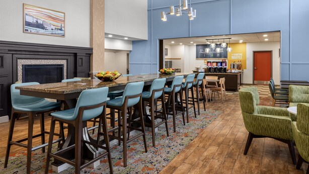 A modern hotel lobby with a long bar area, seating, a fireplace, and a kitchen visible through an open door.