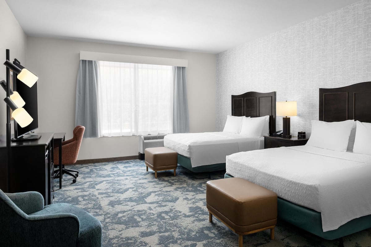 A hotel room with two beds, a desk with a chair, a TV, and a window with curtains.
