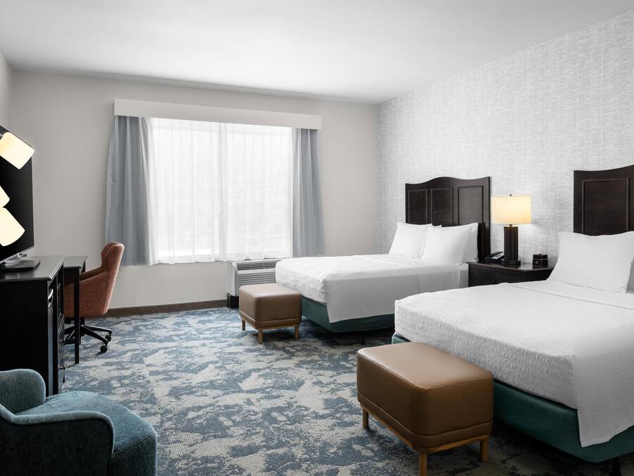 Quarto de hotel com duas camas, uma escrivaninha com cadeira, televisão e janela com cortinas.