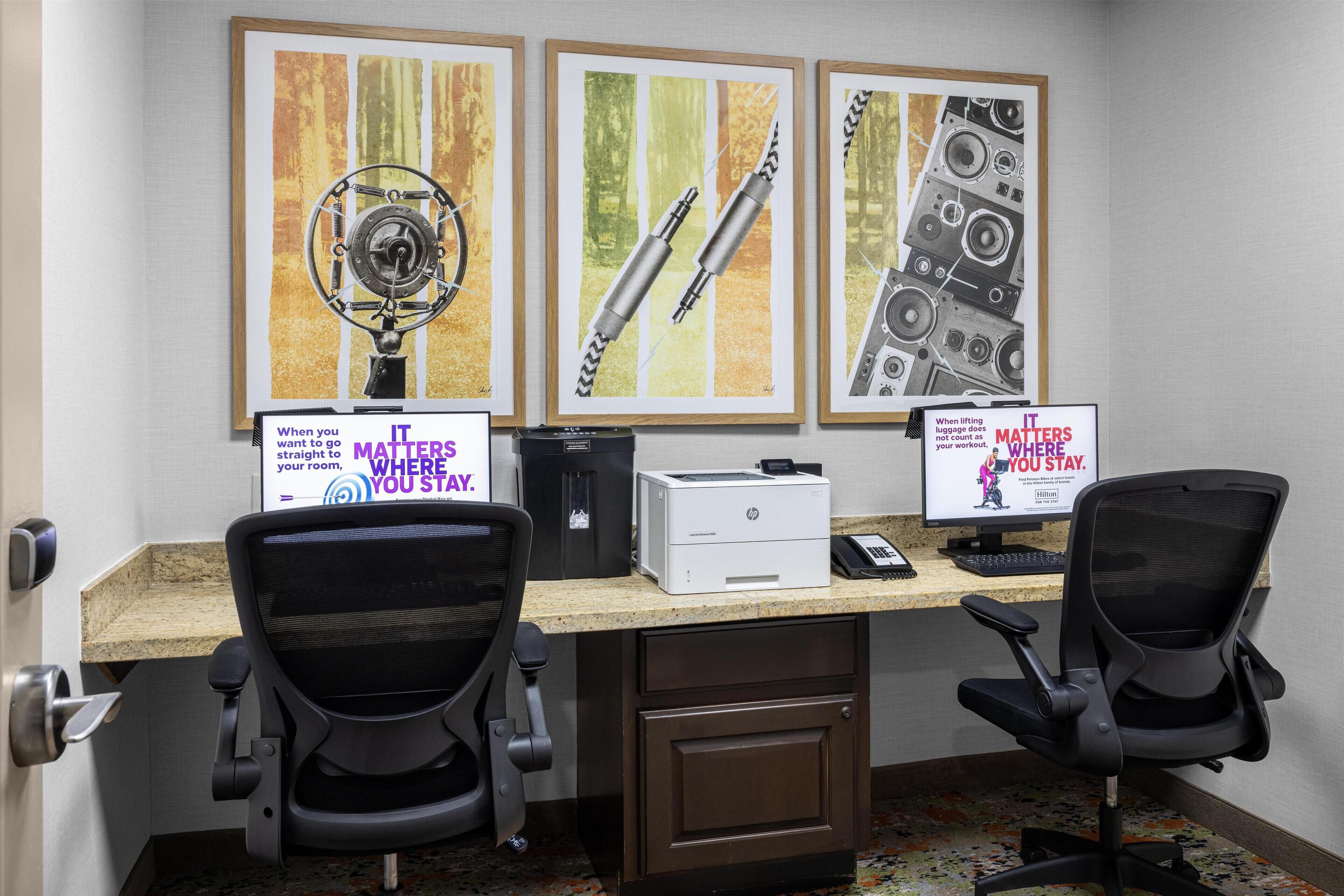 Um espaço de trabalho com duas cadeiras de escritório, uma mesa, dois monitores, uma impressora e quadros na parede.